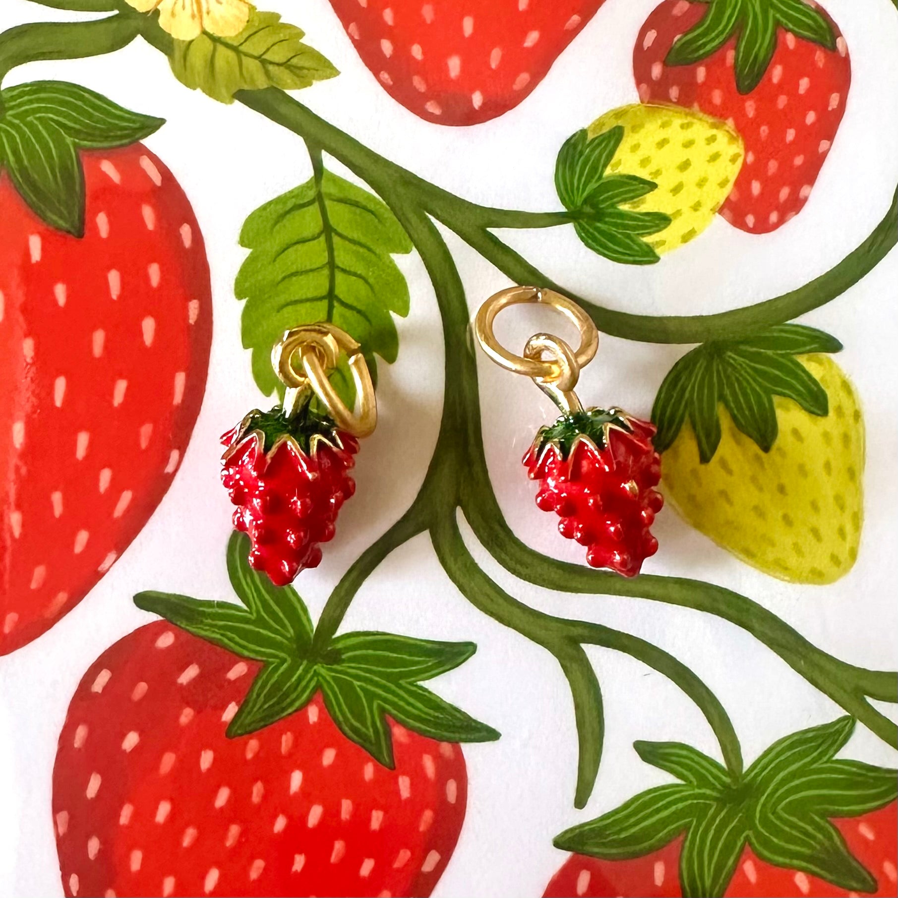 A pair of strawberry cluster drop earrings with gold-tone rings, displayed on a white background printed with a colorful strawberry and leaf pattern.