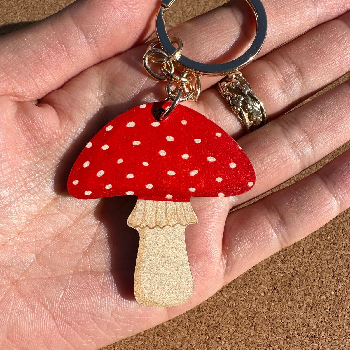 A mushroom-shaped keychain held in a hand. The keychain features a red faux-leather cap with white polka dots and a natural light wood stem, attached to gold-tone metal hardware.