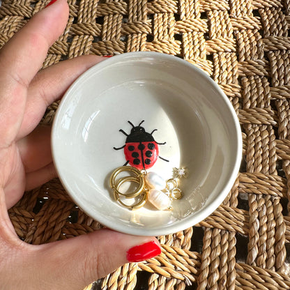 White bowl with a ladybug design containing jewelry on a woven surface