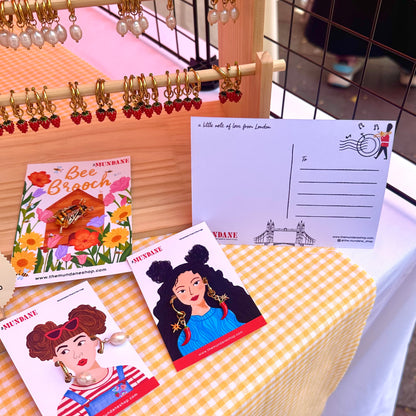 A market stall display featuring handmade earrings with red strawberry charms on wooden hangers, a postcard reading 'a little cell of love from London', and illustrated postcards from 'Bee Brooch'.