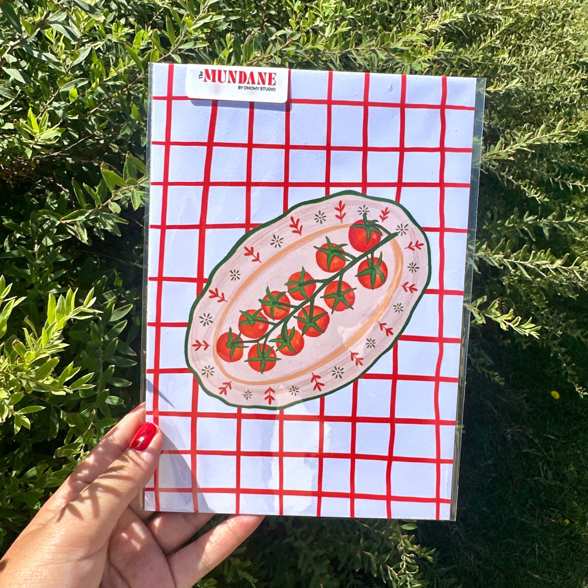 Notebook with tomato design on a red and white checkered cover held in front of green foliage.