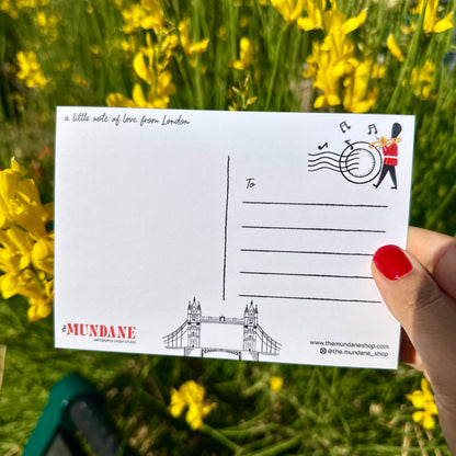 A hand with red nail polish holds a white postcard featuring London-themed illustrations, including Tower Bridge and a postman, against a blurred background of yellow flowers and green grass.