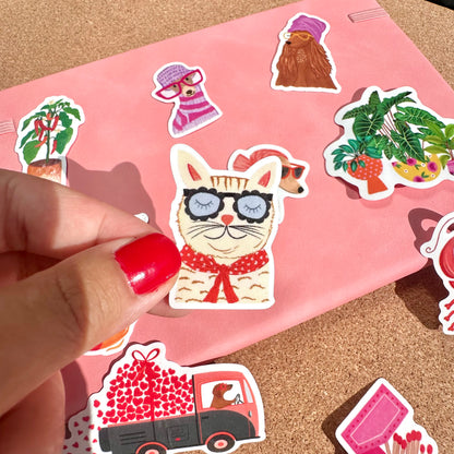 Hand holding a cat sticker with sunglasses on a pink background with other stickers.