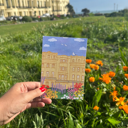 Hand holding a postcard with a building illustration in a garden setting