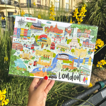A hand with red nail polish holds a clear plastic sleeve containing a London-themed illustrated postcard or art print. The card features colorful drawings of London landmarks, buses, and scenes, with the word 'LONDON' at the bottom.