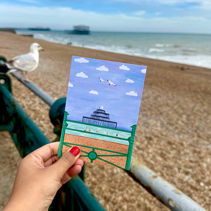 Hand holding a postcard with a beach scene at a seaside, with a seagull on a railing in the background.
