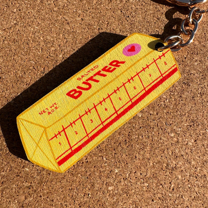 A novelty keychain shaped like a butter carton, featuring a yellow body with red 'BUTTER' text and measurement markings, attached to a silver metal chain. The keychain is photographed on a brown cork surface.