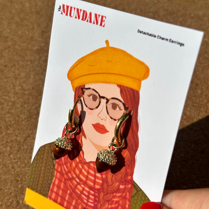 A hand holding a white card featuring an illustrated fashion portrait with the text 'The MUNDANE' and 'Detachable Charm Earrings.' The card shows gold-toned hoop earrings with dangling charm elements, and the background is a brown textured surface.