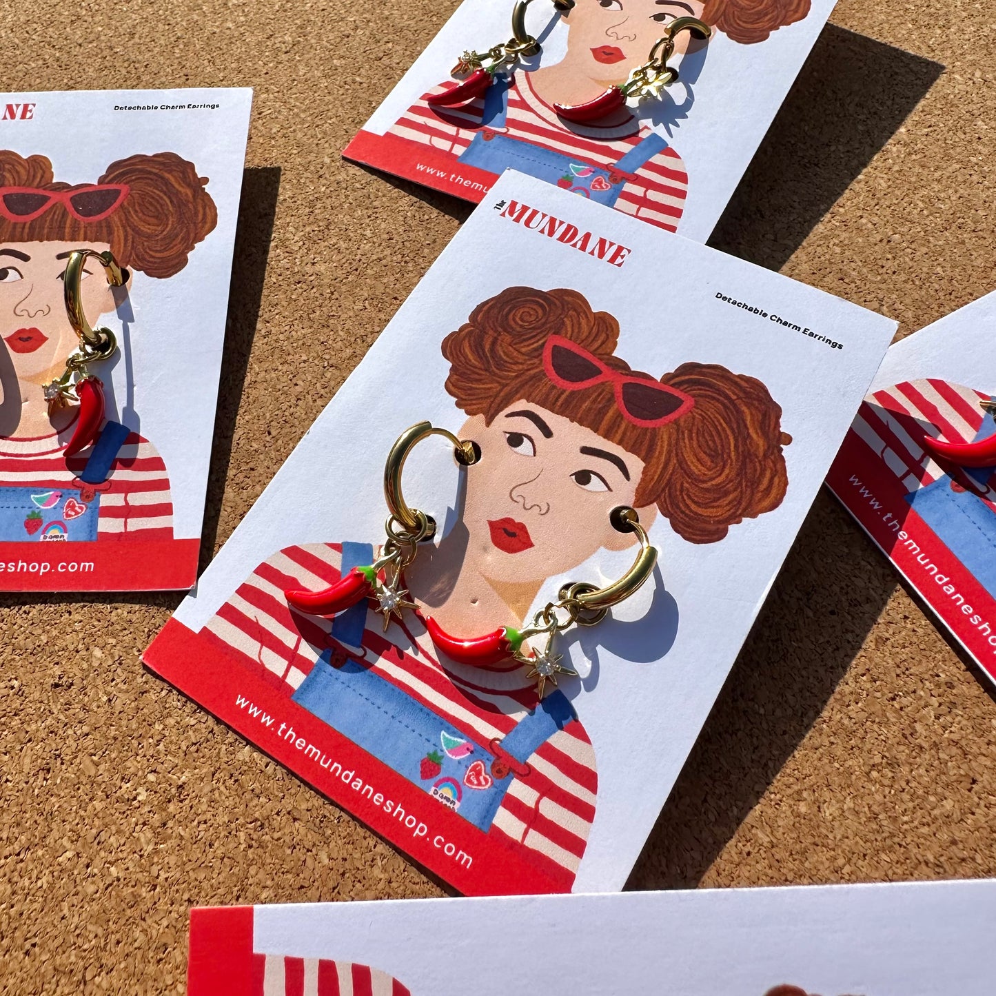 A pair of gold hoop earrings with red and blue dangling charms displayed on a printed card featuring an illustrated woman in a red-and-white striped top. Multiple cards are arranged on a cork surface.