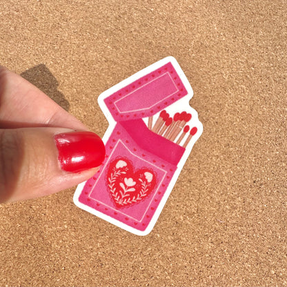 Sticker of a pink matches box with red hearts held by a hand on a cork background
