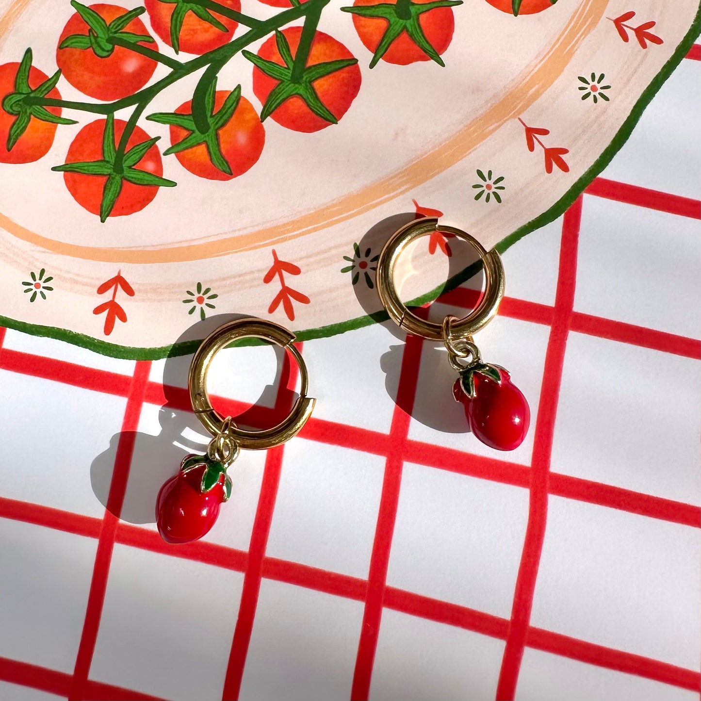 Gold earrings with red tomato charms on a red and white checkered surface.