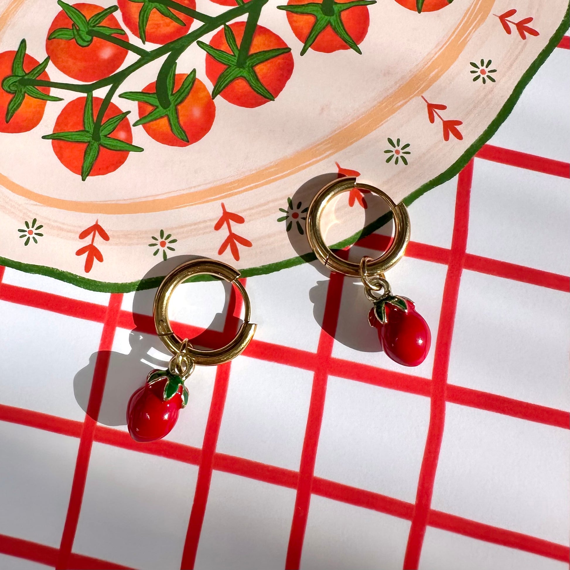 Gold earrings with red tomato charms on a red and white checkered surface.