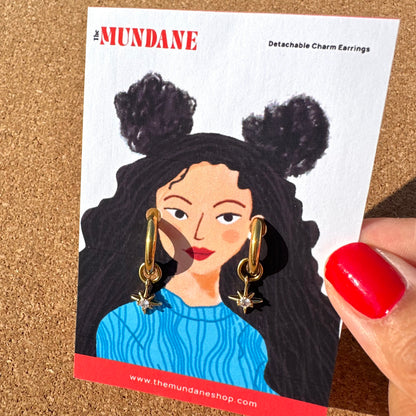 A person's hand with red nail polish holds a white display card featuring an illustration of a woman with black hair in two buns wearing gold huggie earrings with dangling starbursts. The card is branded 'The MUNDANE' and labeled 'Detachable Charm Earrings'.