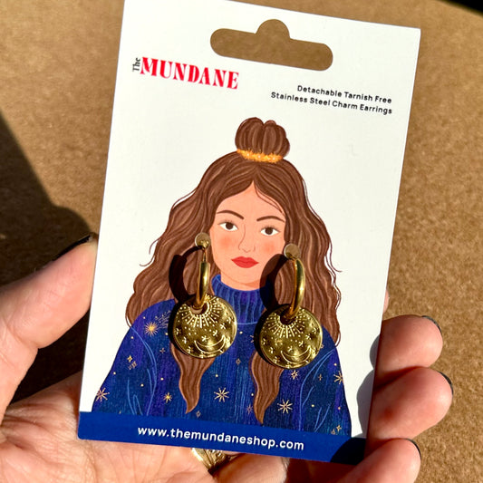 A hand holding a card displaying a pair of gold-toned dangling earrings with circular textured charms. The card has an illustration of a woman wearing the earrings and the brand name 'The Mundane'.