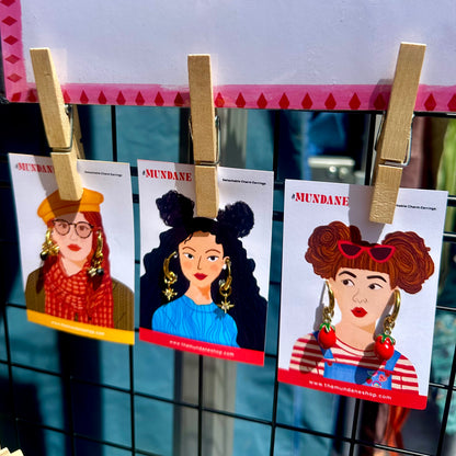 Three printed cards featuring illustrated women hang from a black wire grid with wooden clothespins. The cards have the brand name MUNDANE at the top and colorful portraits with different hair, glasses, jewelry, and outfits.