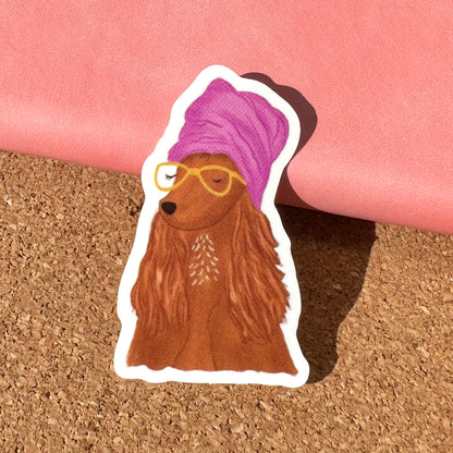 Sticker of a dog with a purple towel on its head and yellow glasses, held against a pink and cork background.