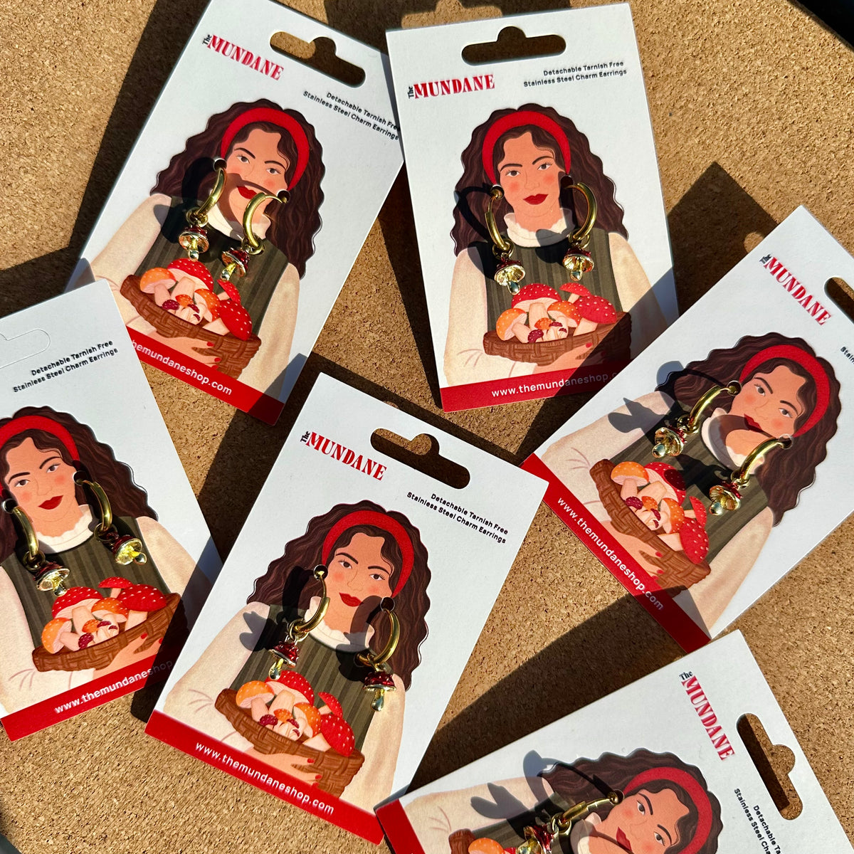 Several Amunave earrings on their original retail backing cards, arranged on a cork surface. The earrings are gold-plated with a red headband, green vest, and a strawberry-themed pendant.