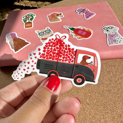 Hand holding a sticker of a red truck with hearts and a dog, with other stickers on a pink background.