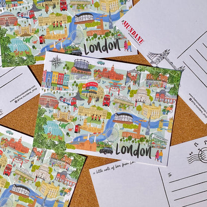 A London-themed illustrated postcard laid on top of several other postcards and a cork board. The main postcard features a colorful cartoon map of London landmarks, rivers, buses, and people, with the word 'London' printed prominently.