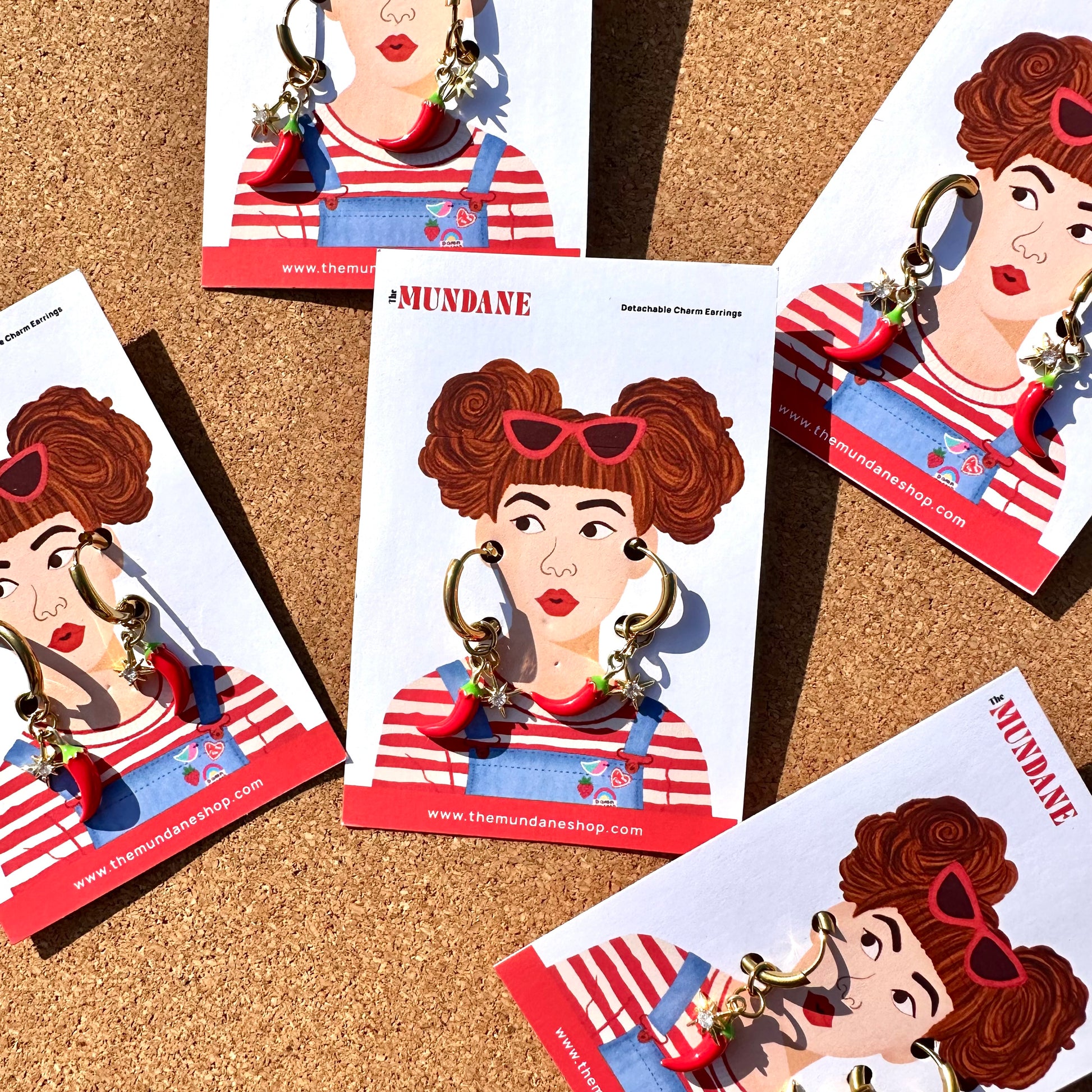 Several printed display cards featuring an illustrated woman wearing red-and-white striped overalls and large hoop earrings, arranged on a cork surface. The cards show the brand name MUNDANE and a website, and the earrings are visible on the cards.
