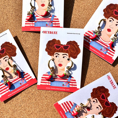Several printed display cards featuring an illustrated woman wearing red-and-white striped overalls and large hoop earrings, arranged on a cork surface. The cards show the brand name MUNDANE and a website, and the earrings are visible on the cards.