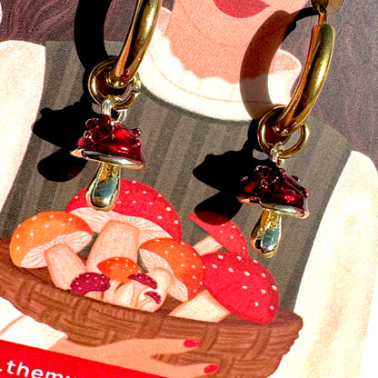 Gold-tone hoop earrings with dangling mushroom charms, including red and orange mushrooms in a basket, shown against a stylized illustrated background.
