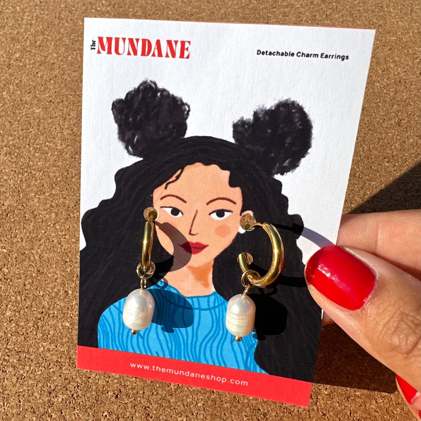 A hand with red nail polish holds a white display card featuring an illustration of a woman wearing gold huggie hoop earrings with white pearl drops. The card is branded 'The MUNDANE' and is photographed against a cork board background.

