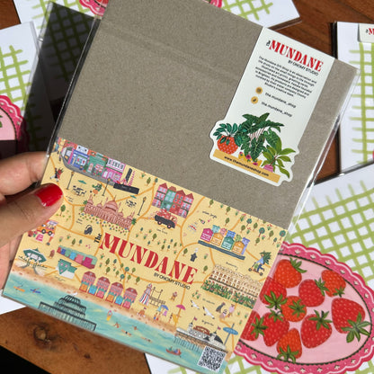 Notebook with 'Mundane' branding held by a hand, surrounded by other items on a wooden surface.