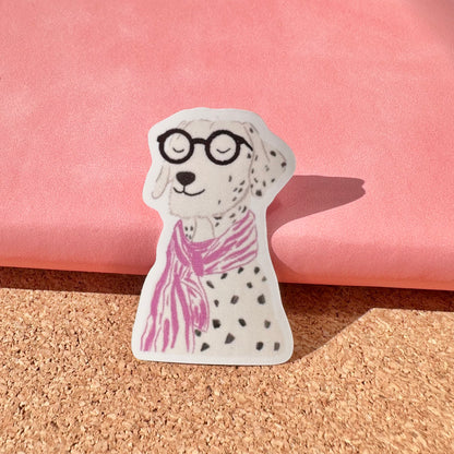 Sticker of a dalmatian dog wearing glasses and a pink scarf on a cork surface with a pink background.