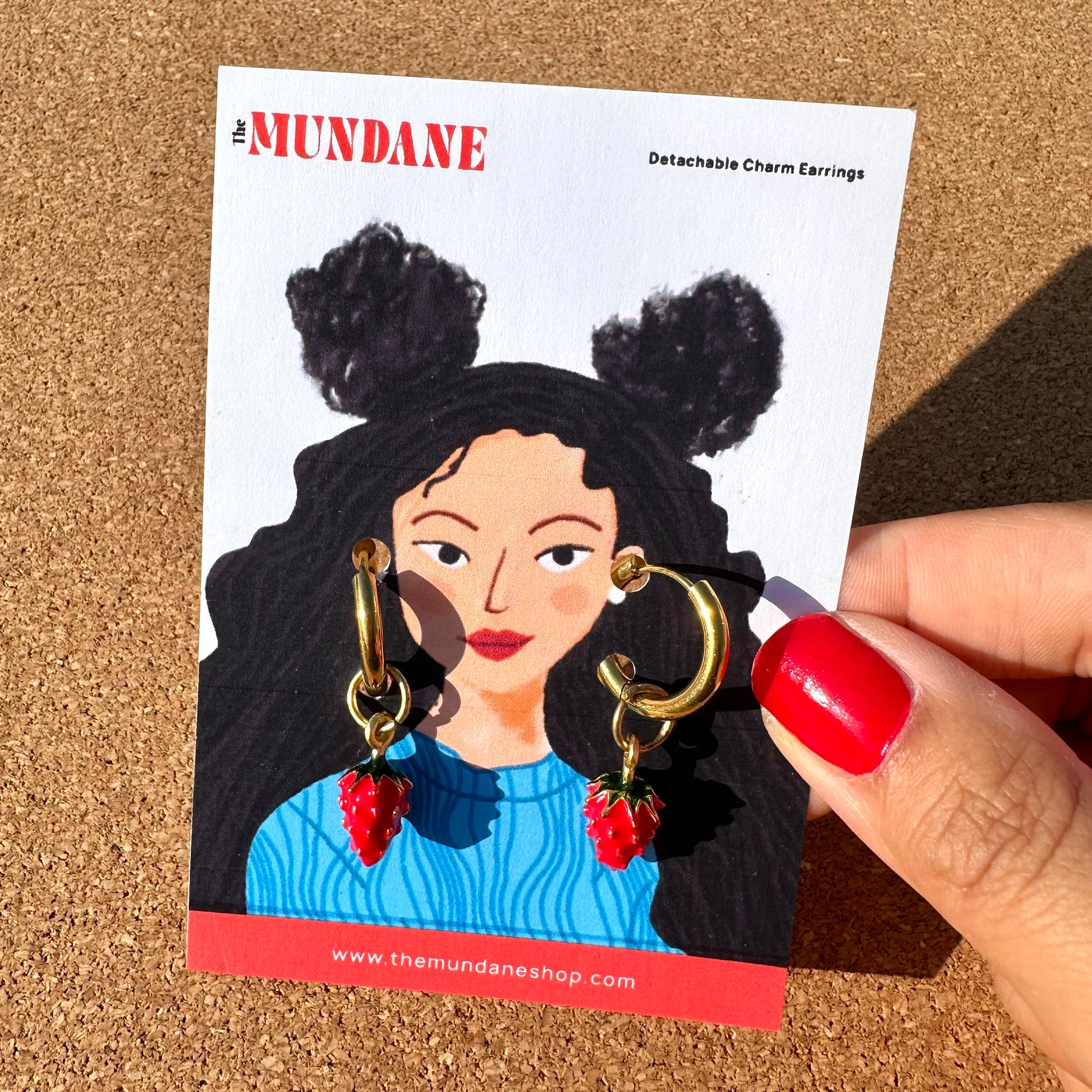 A hand with red nail polish holds a card displaying gold-tone huggie hoop earrings with dangling strawberry charms. The card is branded 'The MUNDANE' and features an illustrated portrait of a woman with black buns.