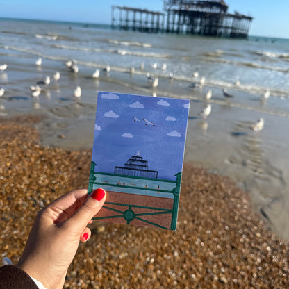 Hand holding a small card with a design of a bench and dome on a beach with a pier in the background.