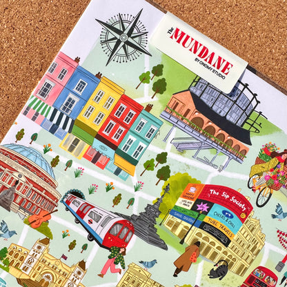 A colorful illustrated map-style print titled "The MUNDANE BY ORNEY STUDIO" laid on a cork surface. The artwork features pastel and bright buildings, trees, a red bus, a bicycle with flowers, people, and street signs in a whimsical city scene.