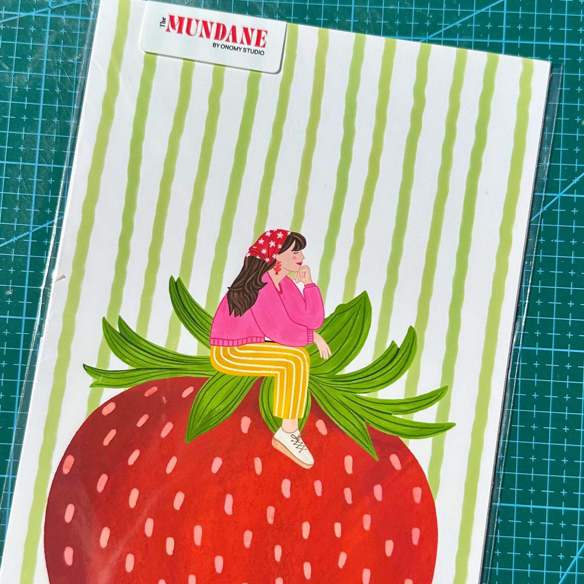 Illustration of a person sitting on a strawberry with green leaves on a striped background, featuring the brand 'Mundane'.