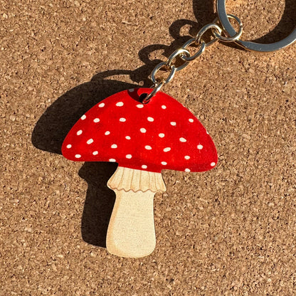 A mushroom-shaped keychain with a red cap covered in white polka dots and a natural wood stem, attached to a silver metal keyring and photographed on a cork surface.