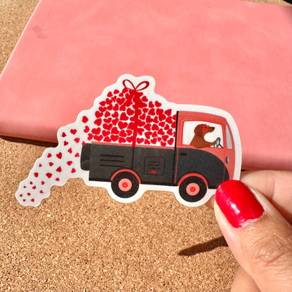 Sticker of a truck filled with hearts held by a hand on a cork surface.