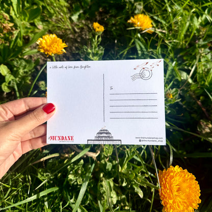 Hand holding a postcard with 'Mundane' branding amidst yellow flowers and green grass