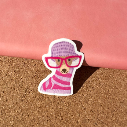 Sticker of a bear wearing a pink hat and glasses on a cork surface.
