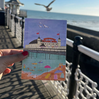 Hand holding a postcard with a beach scene and pier illustration, with a pier and ocean in the background.