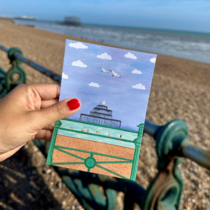 Hand holding a postcard with a beach scene on a pebble beach.
