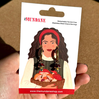A hand holding a white card backing for a pair of dangling earrings. The card has a cartoon illustration of a woman with dark hair, a red headband, and a white top holding a basket of red-and-white mushrooms.