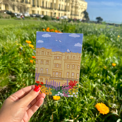 Hand holding a small painting of a building with flowers in a grassy field