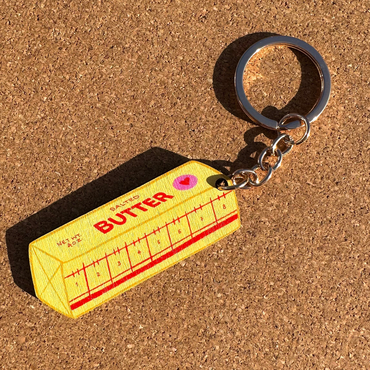 A novelty keychain shaped like a butter carton, featuring a yellow body with red text and measurement markings, attached to a silver metal ring, shown on a cork background.