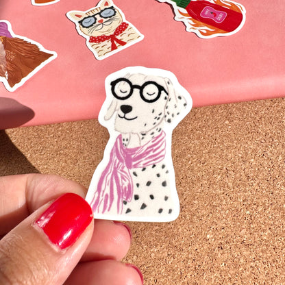 Sticker of a dog wearing glasses and a pink scarf held by a hand on a cork surface with other stickers in the background.