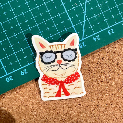 Cat-shaped pin with sunglasses and a red bow tie on a cork surface