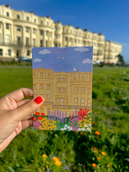 Hand holding a postcard with a building and garden illustration against a blurred outdoor background.