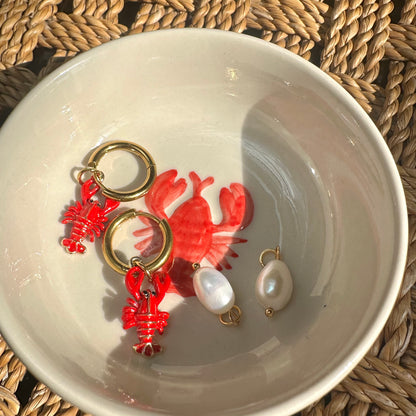 Red lobster-shaped earrings with gold loops and pearl drops on a white dish.