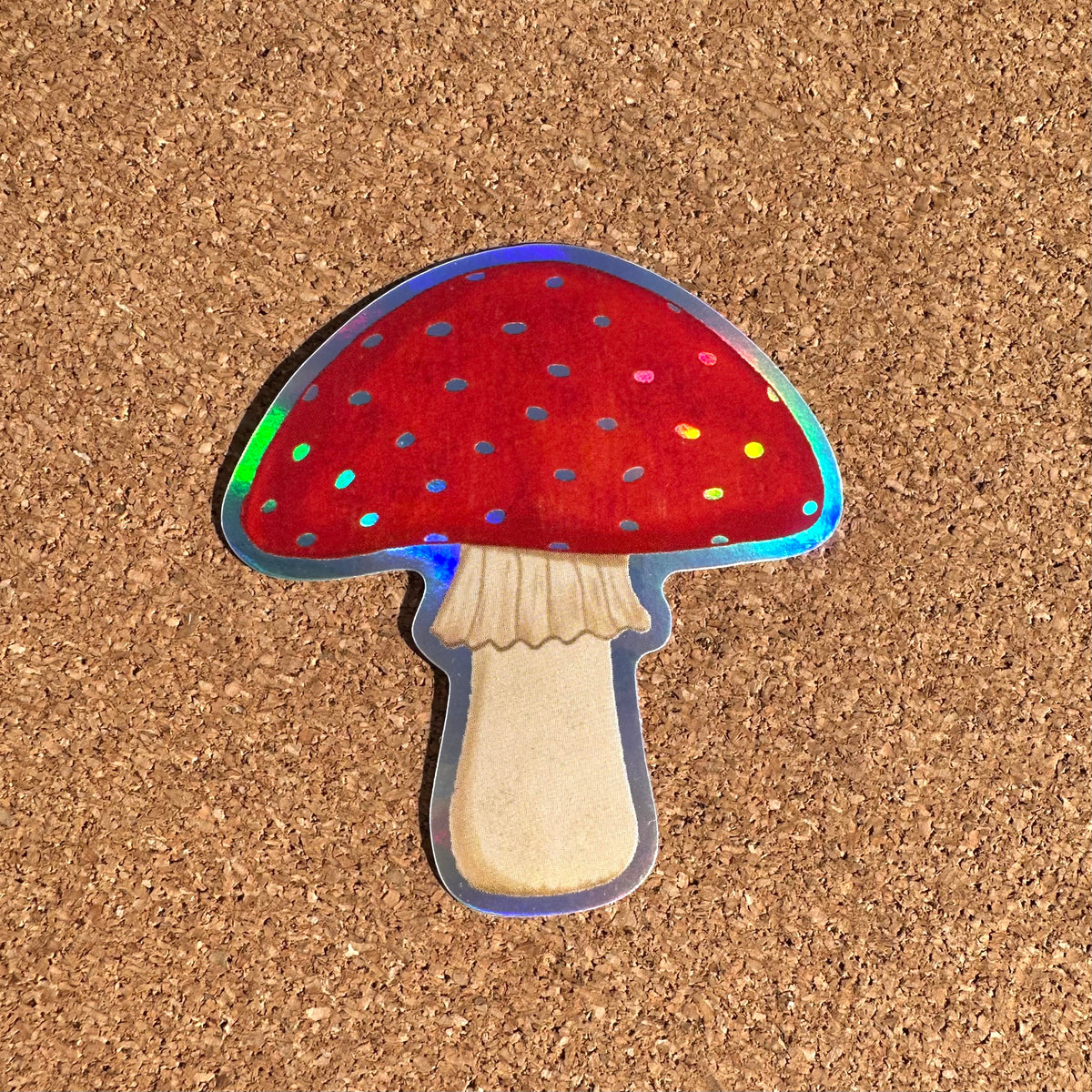A die-cut sticker of a red mushroom with a beige stem and holographic glitter details, placed on a brown cork background.