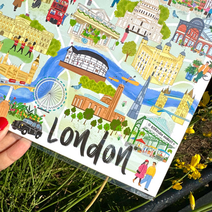 A hand holds a square illustrated London map or postcard-style print outdoors, with London landmarks, a river, trees, and the large word 'London' printed across the front. Yellow flowers and green grass are visible around it.