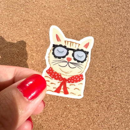 Cat sticker with sunglasses and a red bow held by a hand on a cork background