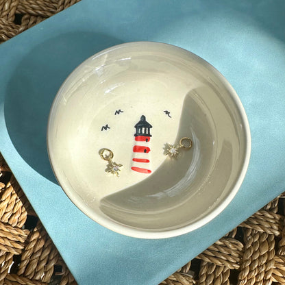 Ceramic bowl with lighthouse design containing jewelry on a textured surface